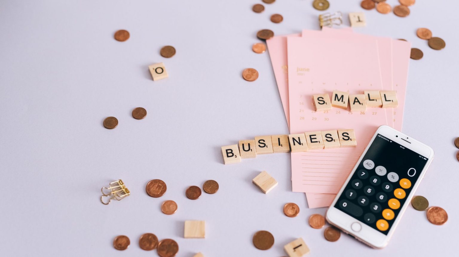 Moedas, calendário e celular com calculadora sobre uma mesa, ilustrando planejamento financeiro para pequenos negócios.
