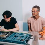 Dois jovens usando óculos de proteção trabalham em um projeto com ferramentas dentro de uma oficina.