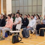 Grupo diverso de adultos acompanha atentamente uma conferência em ambiente interno.