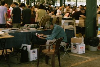 Homem lendo uma revista em meio a barracas de um mercado de livros ao ar livre.