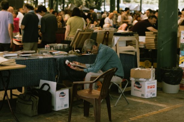 Homem lendo uma revista em meio a barracas de um mercado de livros ao ar livre.