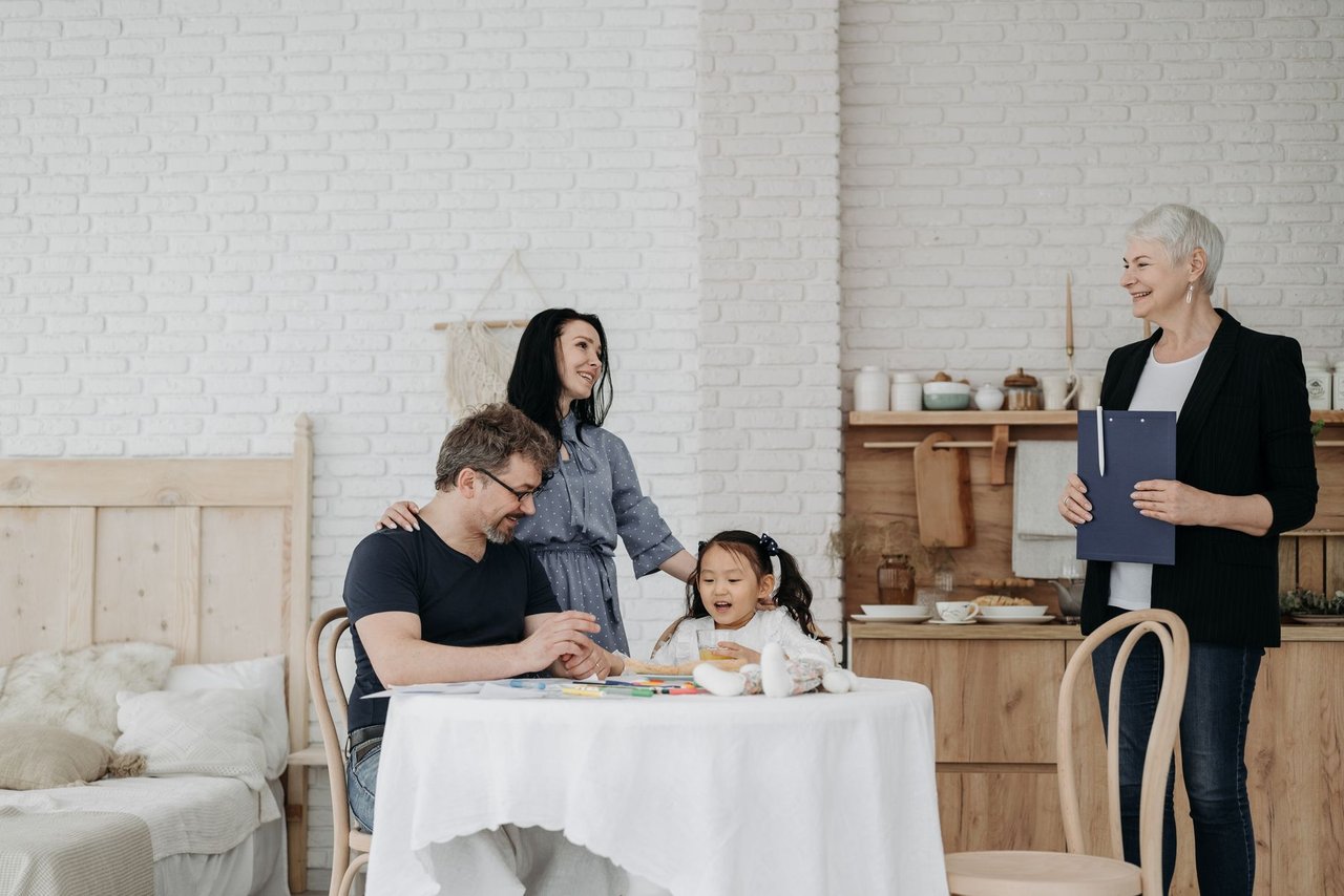 Família conversa à mesa com uma profissional da assistência social em ambiente doméstico.