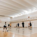 Atletas jogam vôlei em um ginásio esportivo coberto com piso de madeira.