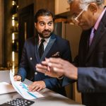 Dois homens analisam documentos financeiros com gráficos durante uma reunião em ambiente interno.
