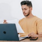 Estudante lê documentos enquanto consulta informações em um laptop.