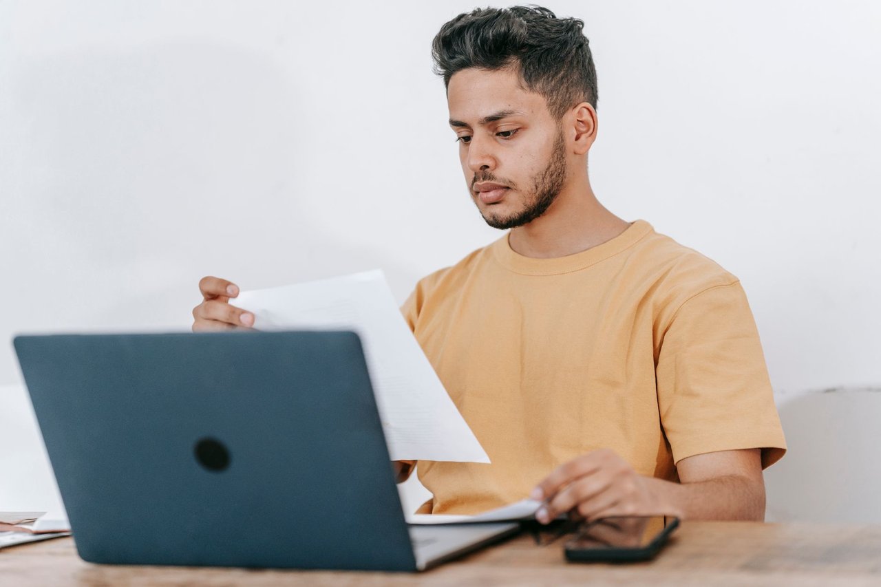 Estudante lê documentos enquanto consulta informações em um laptop.