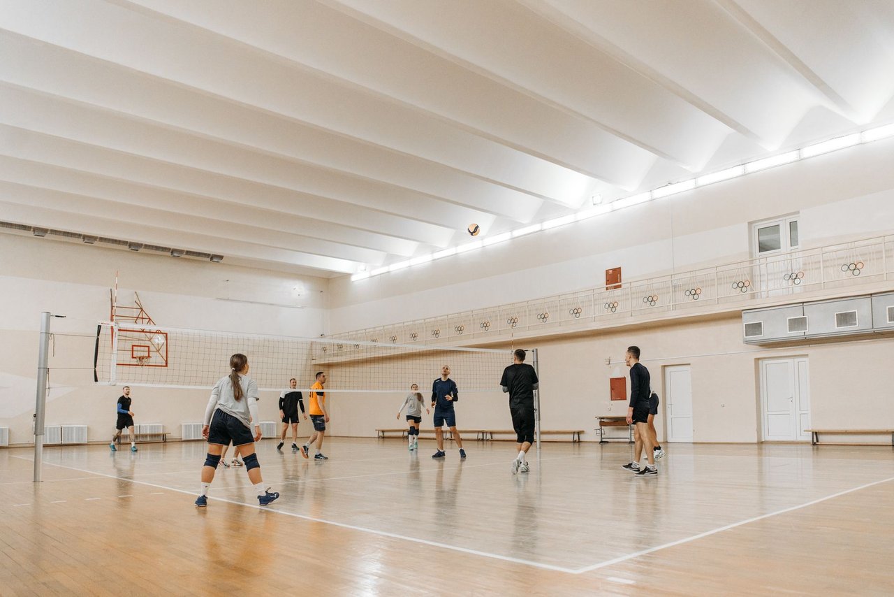 Atletas jogam vôlei em um ginásio esportivo coberto com piso de madeira.