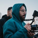 Pessoa opera uma câmera de vídeo ao ar livre, com um monitor acoplado ao equipamento.
